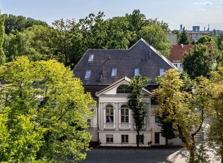 Budynek jest otoczony zielenią, obok znajduje się też sklep spożywczy